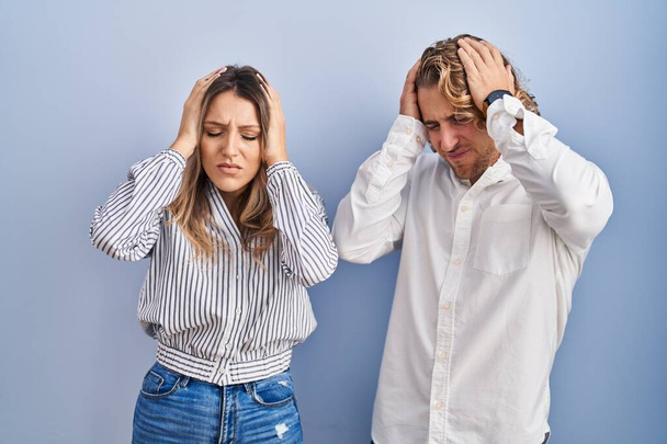 Молодая пара, стоящая на синем фоне, страдает от головной боли в отчаянии и стрессе из-за боли и мигрени. руки на голове.  - Фото, изображение