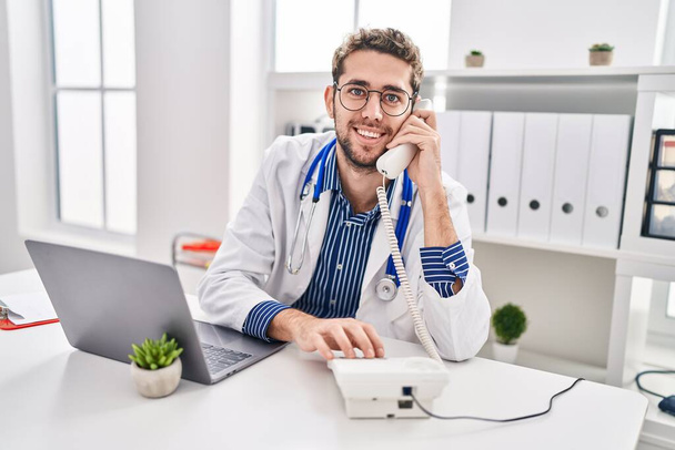 Νεαρός ιατρός που χρησιμοποιεί φορητό υπολογιστή μιλώντας στο τηλέφωνο στην κλινική - Φωτογραφία, εικόνα