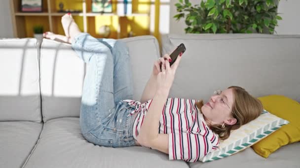 Молодая блондинка с помощью смартфона лежит на диване дома - Кадры, видео