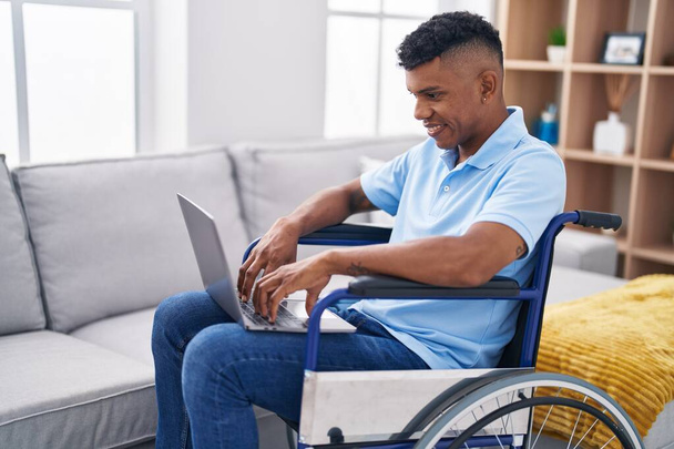 Jeune homme latin utilisant un ordinateur portable assis sur fauteuil roulant à la maison - Photo, image