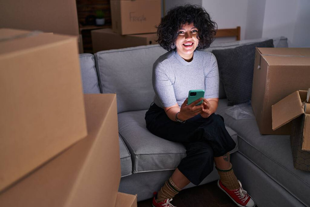 Giovane bella donna ispanica utilizzando smartphone seduto sul pavimento di una nuova casa - Foto, immagini