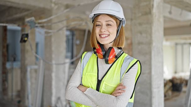 Joven mujer rubia arquitecta sonriendo confiada de pie con los brazos cruzados gesto en el sitio de construcción - Foto, imagen