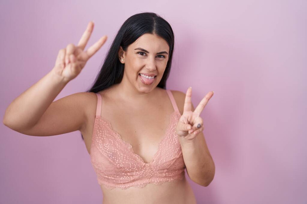 Młoda Latynoska kobieta w różowym staniku uśmiechnięta językiem pokazująca palce obu rąk robiące znak zwycięstwa. numer dwa..  - Zdjęcie, obraz
