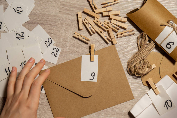 Unrecognizable young woman sticks number on craft bag, fastens with clothespin. Female making kraft paper for homemade advent calendar Made with your own hands step by step DIY crafts do it yourself - Fotó, kép