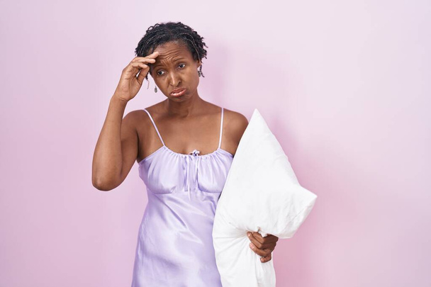 African woman with dreadlocks wearing pajama hugging pillow worried and stressed about a problem with hand on forehead, nervous and anxious for crisis  - Foto, Imagen