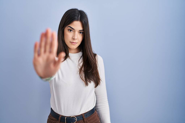 Young brunette woman standing over blue background doing stop sing with palm of the hand. warning expression with negative and serious gesture on the face.  - Foto, Imagen