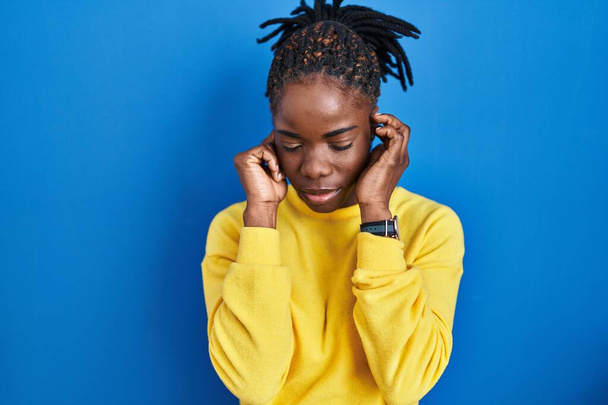 Beautiful black woman standing over blue background covering ears with fingers with annoyed expression for the noise of loud music. deaf concept.  - Foto, Imagen