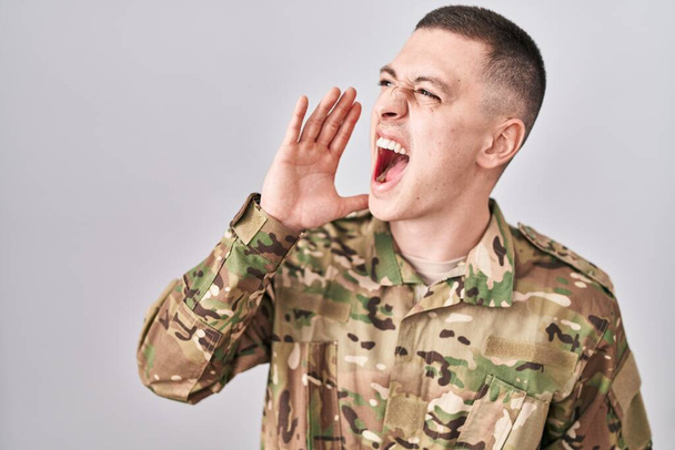 Jeune homme portant un uniforme de camouflage de l'armée criant et criant haut et fort à côté avec la main sur la bouche. concept de communication.  - Photo, image
