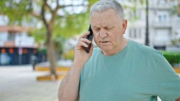 Middelbare leeftijd grijsharige man aan het praten op smartphone met serieuze expressie in het park - Foto, afbeelding