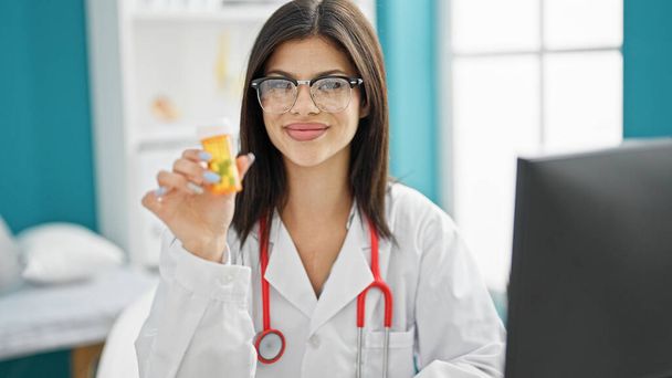 Genç beyaz kadın doktor elinde ilaç şişesiyle klinikte gülümsüyor. - Fotoğraf, Görsel