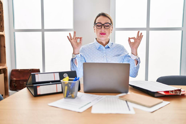 Young hispanic woman working at the office wearing glasses relax and smiling with eyes closed doing meditation gesture with fingers. yoga concept.  - Foto, Imagem