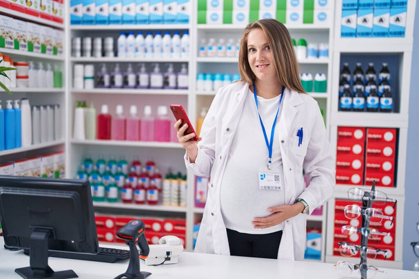 Fiatal terhes nő gyógyszerész okostelefont használ a gyógyszertárban - Fotó, kép