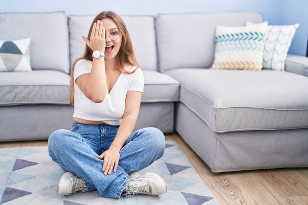 Young caucasian woman sitting on the floor at the living room covering one eye with hand, confident smile on face and surprise emotion.  - Фото, изображение