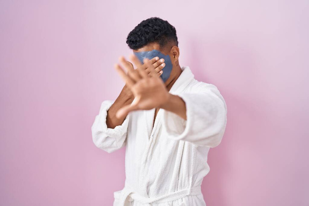 Young hispanic man wearing beauty face mask and bath robe covering eyes with hands and doing stop gesture with sad and fear expression. embarrassed and negative concept.  - Photo, image