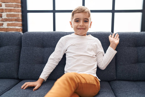 Αξιολάτρευτο ισπανόφωνο αγόρι λέει γεια με το χέρι κάθεται στον καναπέ στο σπίτι - Φωτογραφία, εικόνα
