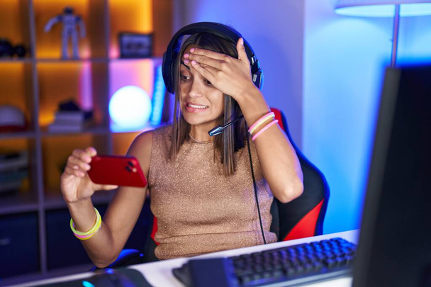 Νεαρή ισπανόφωνη γυναίκα που παίζει βιντεοπαιχνίδια με smartphone αγχωμένη και απογοητευμένη με το χέρι στο κεφάλι, έκπληκτη και θυμωμένη  - Φωτογραφία, εικόνα