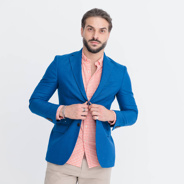portrait of sexy unshaved man looking forward while buttoning blue jacket and posing in front of light grey background - Φωτογραφία, εικόνα