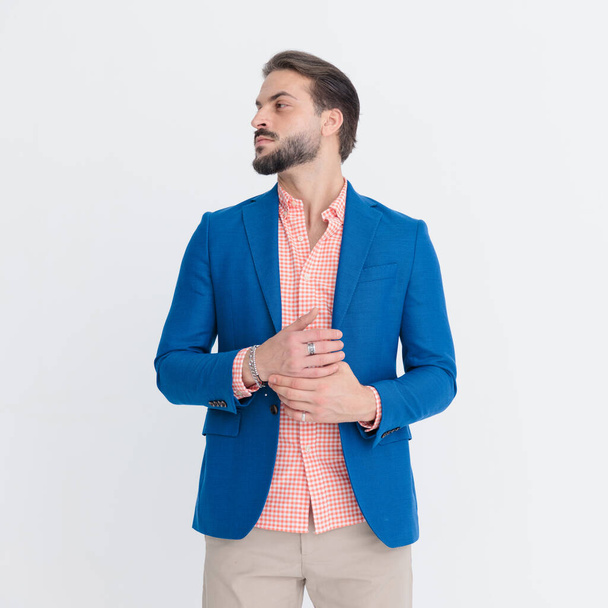 smart casual νεαρός άνδρας με μπλε σακάκι με καρό πουκάμισο που αγγίζει τα δάχτυλα και κοιτάζει προς τα πλάγια μπροστά από ανοιχτό γκρι φόντο στο στούντιο - Φωτογραφία, εικόνα