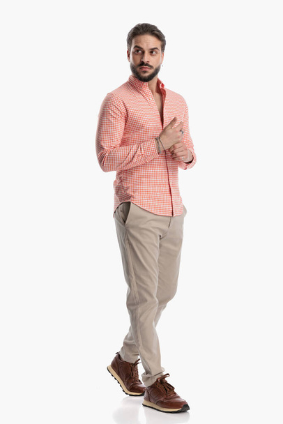 full body picture of attractive young man in plaid shirt looking to side and adjusting bracelets while walking in front of white background in studio - Photo, Image