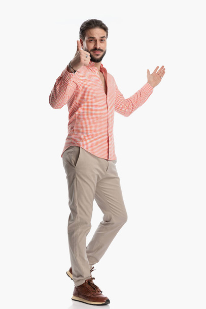 confident young man in plaid shirt making thumbs up sign while showing to side with other hand and walking in front of white background - Photo, image