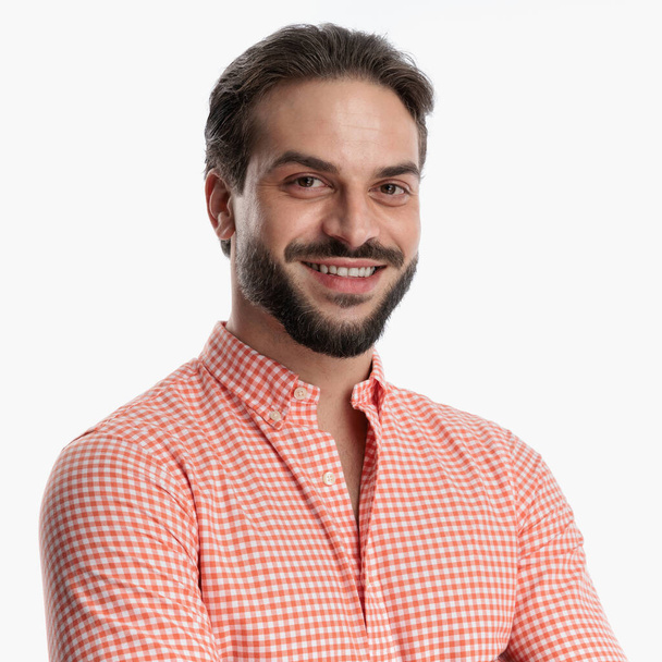 portrét šťastného mladého muže s vousy v kostkované košili a s úsměvem při sebevědomém pózování před bílým pozadím ve studiu - Fotografie, Obrázek