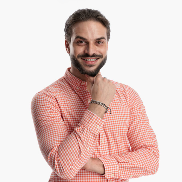 portrait of happy casual guy in plaid shirt touching chin and thinking while folding arms and smiling in front of white background in studio - Photo, Image