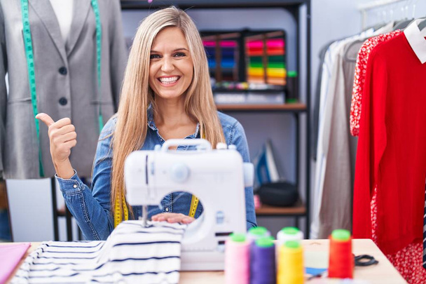 Mujer rubia modista diseñador utilizando máquina de coser que apunta a la parte posterior con la mano y los pulgares hacia arriba, sonriendo confiado  - Foto, Imagen