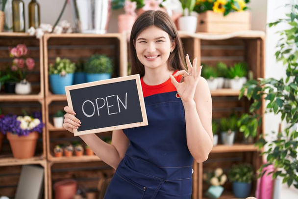 Joven mujer caucásica trabajando en floristería sosteniendo letrero abierto haciendo signo de ok con los dedos, sonriendo gesto amistoso excelente símbolo  - Foto, imagen