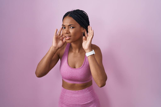 Afrikkalainen amerikkalainen nainen punokset yllään urheiluvaatteet vaaleanpunainen tausta yrittää kuulla molemmat kädet korva ele, utelias juoruja. kuulo-ongelma, kuuro  - Valokuva, kuva