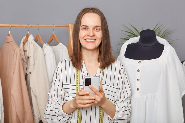 Attractive positive charming woman seamstress standing near new dress on mannequin against gray wall and hangers with clothes holding mobile phone looking at camera. - 写真・画像