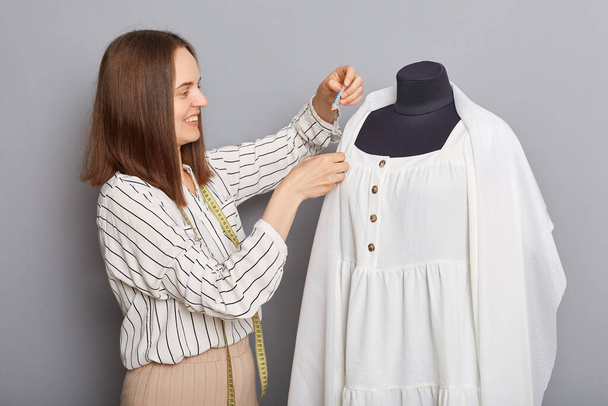 Entrepreneur's clothing brand. Skilled tailor's expertise. Dark haired young woman standing near new dress on mannequin iisolated over gray background. - Foto, Imagem