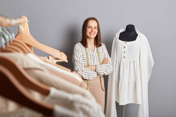 Unique garments, happy customers. Attractive smiling woman tailor standing near new dress on mannequin iisolated over gray background keeps hands crossed looking at camera. - Фото, изображение