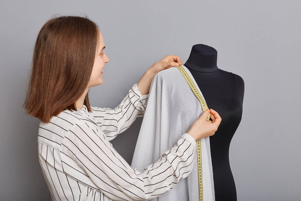 Entrepreneurial clothing venture. Custom garment creation. Cloth and textile production. Smiling woman needlewoman standing near new dress on mannequin iisolated over gray background. - Foto, Imagen