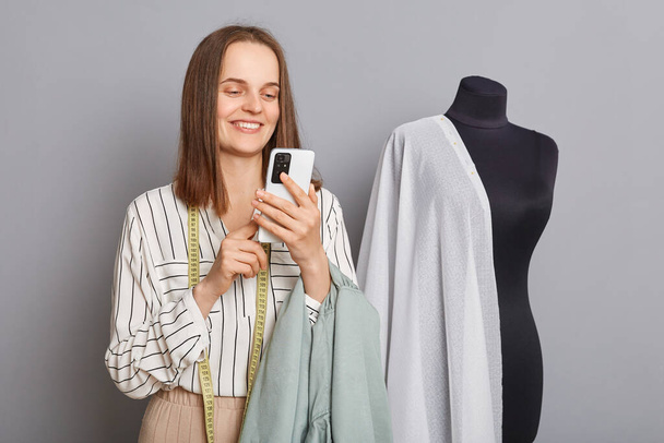 Cute charming positive woman needlewoman standing near new dress on mannequin iisolated over gray background using mobile phone browsing internet finds new ideas. - Zdjęcie, obraz