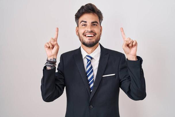 Fiatal spanyol férfi tetoválásokkal, öltönyben és nyakkendőben, mosolyogva, csodálkozva és meglepve, ujjal mutogatva, felemelt karral..  - Fotó, kép