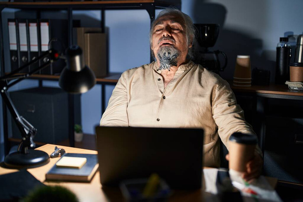 Középkorú férfi ősz hajjal dolgozik az irodában éjszaka álmos és fáradt, kimerült a fáradtság és a másnaposság, lusta szemek reggel.  - Fotó, kép