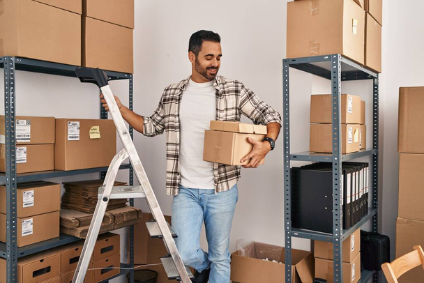 Young hispanic man ecommerce business worker holding packages standing at office - Фото, изображение