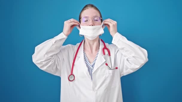 Νεαρή ξανθιά γυναίκα γιατρός φορώντας ιατρική μάσκα κάνει χειρονομία επάνω σε απομονωμένο μπλε φόντο - Πλάνα, βίντεο
