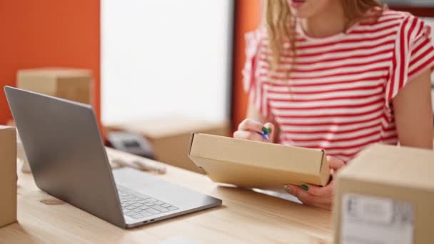 Νεαρή ξανθιά γυναίκα υπάλληλος ηλεκτρονικού εμπορίου χρησιμοποιώντας το laptop γράφοντας στο πακέτο στο γραφείο - Πλάνα, βίντεο