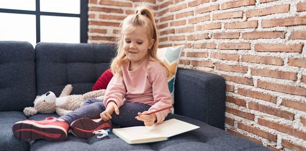 Adorable chica rubia estudiante de preescolar sentado en el sofá dibujo en el cuaderno en casa - Foto, imagen