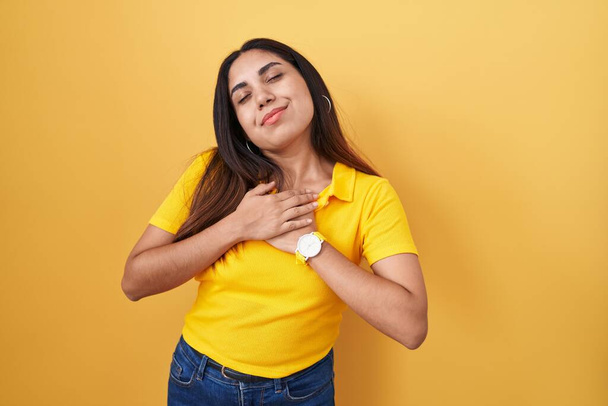 Young arab woman standing over yellow background smiling with hands on chest with closed eyes and grateful gesture on face. health concept.  - Zdjęcie, obraz