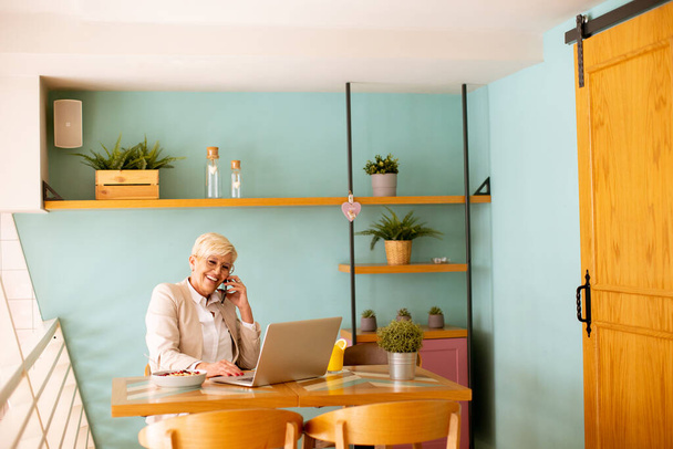 Vanhempi nainen käyttää matkapuhelinta työskennellessään kannettavan tietokoneen kanssa ja juoden tuoretta appelsiinimehua kahvilassa - Valokuva, kuva