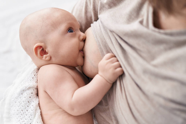 Adorable caucasian baby sitting on bed with mother sucking breast to eat at bedroom - Foto, Imagen