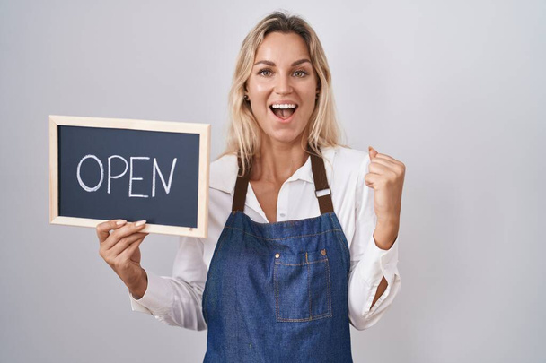 Young blonde woman wearing apron holding blackboard with open word screaming proud, celebrating victory and success very excited with raised arms  - Foto, imagen