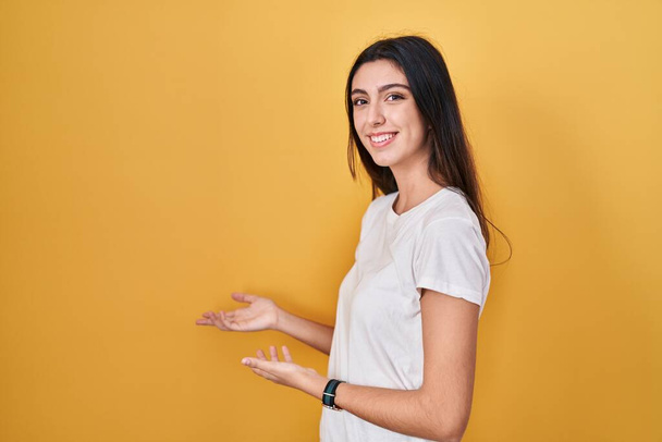 Mladá krásná žena stojící nad žlutým pozadím zvoucí ke vstupu s úsměvem přírodní s otevřenou rukou  - Fotografie, Obrázek