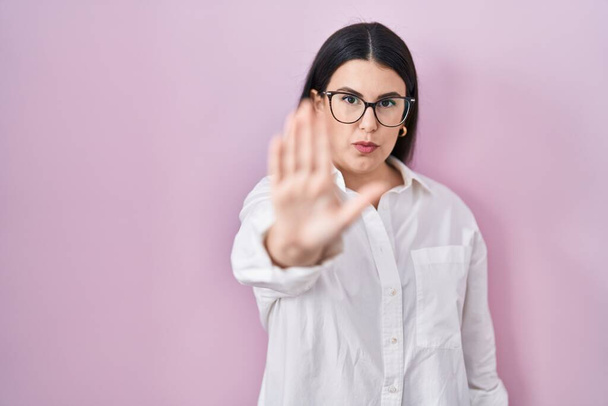 Jonge brunette vrouw staan over roze achtergrond doen stoppen met zingen met palm van de hand. waarschuwingsuitdrukking met negatief en ernstig gebaar op het gezicht.  - Foto, afbeelding