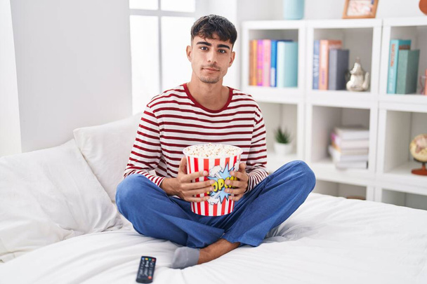 Joven hispano comiendo palomitas de maíz sentado en la cama viendo una película relajada con expresión seria en la cara. simple y natural mirando a la cámara.  - Foto, imagen