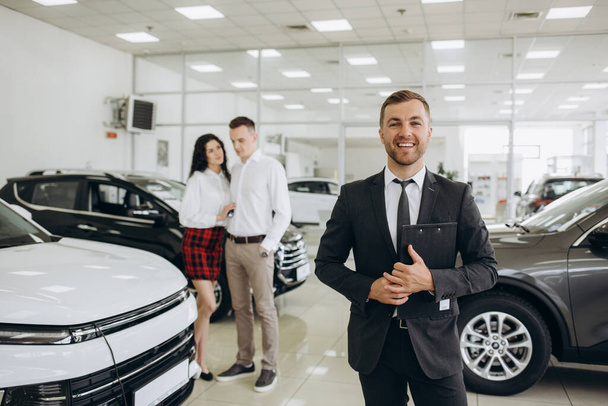 Focused photo of young male shop assistant explaining talking offering buying new car to young caucasian family husband and wife at auto dealer shop - Photo, Image