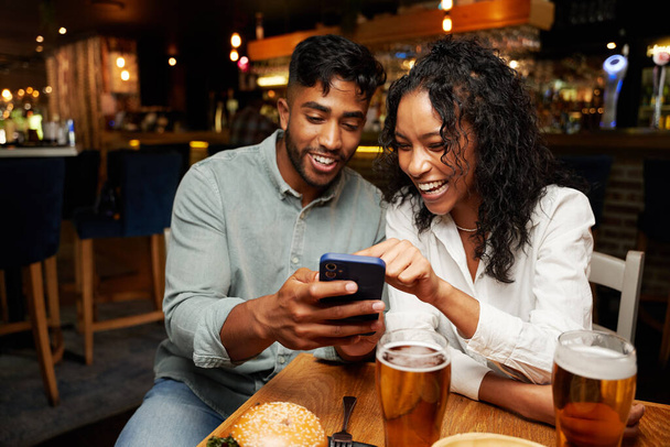 Young multiracial couple wearing casual clothing smiling while typing on mobile phone at restaurant - 写真・画像