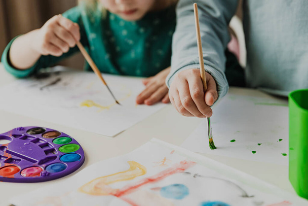 Two cute little sisters, classmates are painting on table. Small preschool and school girl. Kids use brush in kindergarten or school. Adorable preschooler in living room. Home schooling concep - Foto, Imagem